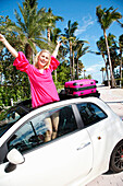 Junge blonde Frau in pinkem Sommerkleid steht in der Dachfensteröffnung eines Wagens