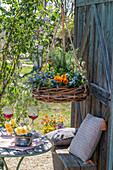 Blumenampel mit Vergissmeinnicht, Hornveilchen (Viola Cornuta), Rosmarin und Tisch mit Narzissen, Osterdeko und Weingläser auf Terrasse