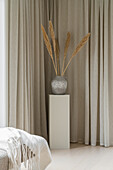 Standing area with silver vase and dried grasses in front of beige curtains