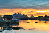 Schneebedeckte Berge und rote Fischerhütten unter dem brennenden Himmel in der Morgendämmerung, Ballstad, Vestvagoy, Lofoten Inseln, Norwegen, Skandinavien, Europa