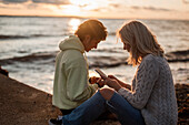 Paar sitzt bei Sonnenuntergang am Strand und benutzt seine Handys
