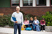 Smiling teacher in front of school, schoolchildren in background