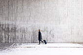 Young man with suitcase walking along wall in winter