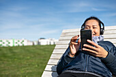 Young woman with smartphone relaxing on wooden sun chair
