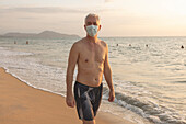 Älterer Mann, der mit einer Gesichtsmaske am Strand spazieren geht