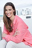 A young woman wearing a salmon-coloured shirt with a pearl necklace and light coloured trousers