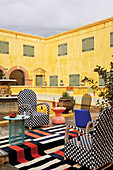 Seating area with checkered armchairs and striped carpet in the courtyard of a castle with bar cart and designer stools in front of a round table