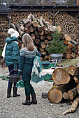 Zwei Frauen in Winterkleidung vor Grillstelle, eine trägt eine Wolldecke und einen grünen Wasserkessel, die andere trägt einen großen Dutch Oven