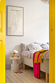 Bedroom with yellow door, light-coloured bed, city map poster and colourful textiles