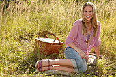 Blonde Frau in rosa Bluse und Jeans-Caprihose mit Picknickkorb auf der Wiese