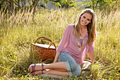 Blonde woman in pink blouse and jeans capri pants with picnic basket on the meadow