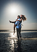 A Man Holds A Woman In The Shallow Water Along The Coast Looking Out At The Ocean; Tarifa, Cadiz, Andalusia, Spain