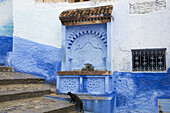 Schwarze Katze geht die Treppe hinauf, vorbei an einem Wasserbrunnen; Chefchaouen, Marokko