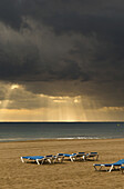 Sonnenstrahlen scheinen durch die dunklen Wolken über dem Ozean und Liegestühle am Strand; Benidorm, Spanien