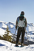 Female Snowshoer Looking Over Snow Covered Mountain Range And Blue Sky; Banff, Alberta, Canada