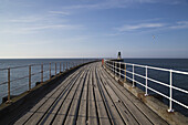 Ein hölzerner Pier mit weißem Geländer, der zum Wasser hinausführt; Whitby, Yorkshire, England