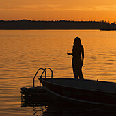 Silhouette einer Frau, die ein Glas Wein in der Hand hält, während sie auf einem Dock an einem goldenen See bei Sonnenuntergang steht; Lake Of The Woods, Ontario, Kanada