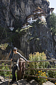 Eine Frau steht an einem Geländer mit dem Taktsang-Palphug-Kloster im Hintergrund; Paro, Bhutan