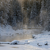 Sunlight Through The Misty Air Over A Snow Covered Landscape And Bridge Crossing Over Water; Emerald Lake, British Columbia, Canada