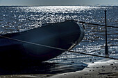 Silhouette eines starren aufblasbaren Zodiacs am Strand mit einem aus dem Sand ragenden Paddel, die Sonne glitzert auf dem arktischen Ozean und ein blauer Himmel im Hintergrund; Spitzbergen, Svalbard, Norwegen