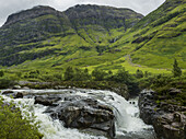 Wasser in einem Fluss, der über Felsen und saftiges Gras in den Hügeln rauscht; Schottland