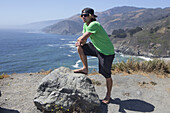 Ein junger Mann steht auf einem Bergrücken mit Blick auf den Ozean und die kalifornische Küste; Big Sur, Kalifornien, Vereinigte Staaten von Amerika