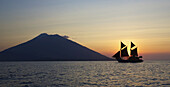 Ocean View With Yacht With Sails And Volcano Silhouetted Against The Setting Sun