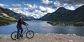 Ein Mann sitzt auf einem Fahrrad am Rande des Wassers mit den Rocky Mountains und einem See im Hintergrund, Waterton Lakes National Park; Alberta, Kanada