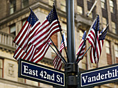 Amerikanische Flaggen an einem Mast über den Straßenschildern an der Kreuzung von East 42nd Street und Vanderbilt; New York City, New York, Vereinigte Staaten von Amerika