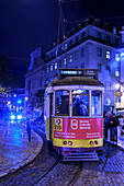 Straßenbahn und Fußgänger in der Abenddämmerung mit Lichtern, die das Straßenleben beleuchten; Lissabon, Region Lisboa, Portugal