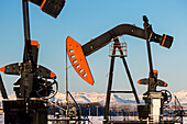 Close-up of pumpjack  in a snow-covered field with snow-covered mountains and blue sky in the background, West of Airdrie; Alberta, Canada