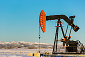 Pumpjack in a snow-covered field with snow-covered mountains and blue sky in the background, West of Airdrie; Alberta, Canada