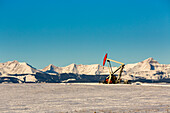 Pumpjack in a snow-covered field with snow-covered mountains and blue sky in the background; Longview, Alberta, Canada