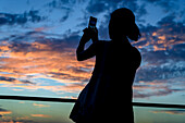 Silhouette einer Frau, die den Sonnenuntergang mit einem Smartphone fotografiert; Havanna, Kuba