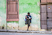 Cuban man stands leaning against the wall of a building along a street; Havana, Cuba