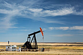 Pumpjack auf einem Feld mit Hügeln, Wolken und blauem Himmel im Hintergrund, westlich von Airdrie; Alberta, Kanada