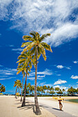 A row of palm trees and a young girl in bikini with surfboard walking past Duke Kahanamoku Lagoon heading to Kahanamoku beach; Oahu, Hawaii, United States of America