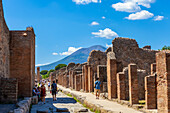 Touristen gehen durch die ausgegrabenen Ruinen auf einer Straße in Pompeji mit dem Vesuv im Hintergrund; Pompeji, Provinz Neapel, Kampanien, Italien