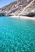 Tourists walking on the beach along the clear, turquoise water of Galazia Nera Bay; Polyaigos Island, Cyclades, Greece