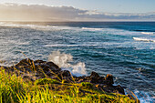 Meereswellen, die bei Sonnenuntergang an die Felsen und das grasbewachsene Ufer rollen, mit einer ruhigen Meereslandschaft und Wolken am Horizont des Ho'okipa Lookout bei Paia; Maui, Hawaii, Vereinigte Staaten von Amerika