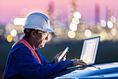 Man working on a laptop and smart phone with an oil refinery in the background; Alberta, Canada