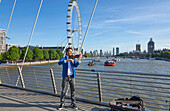 Busker playing violin on a bridge over the River Thames with boats and the London Eye in the background; London, England