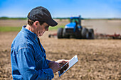 Landwirt nutzt ein Tablet, während er auf einem landwirtschaftlichen Feld steht und ein Traktor mit Geräten das Feld besät; Alberta, Kanada