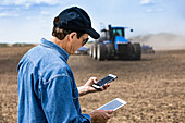 Landwirt benutzt ein Smartphone und ein Tablet, während er auf einem landwirtschaftlichen Feld steht und den Traktor und die Geräte beim Säen des Feldes beobachtet; Alberta, Kanada
