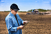 Farmer using a tablet while standing on a farm field and a tractor and equipment seeds the field; Alberta, Canada