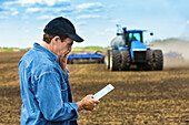 Ein Landwirt benutzt ein Smartphone und ein Tablet, während er auf einem landwirtschaftlichen Feld steht und den Traktor und die Geräte beim Säen des Feldes beobachtet; Alberta, Kanada