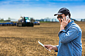 Landwirt benutzt ein Smartphone und ein Tablet, während er auf einem landwirtschaftlichen Feld steht und den Traktor und die Geräte beim Säen des Feldes beobachtet; Alberta, Kanada