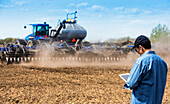Ein Landwirt benutzt ein Tablet, während er auf einem Acker steht und ein Traktor und Geräte das Feld bestellen; Alberta, Kanada