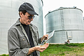 A farmer with a handful of harvested wheat stands with a tablet beside grain bins; Alberta, Canada