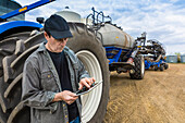 Landwirt nutzt ein Tablet, während er auf einem Bauernhof neben den Geräten steht; Alberta, Kanada
